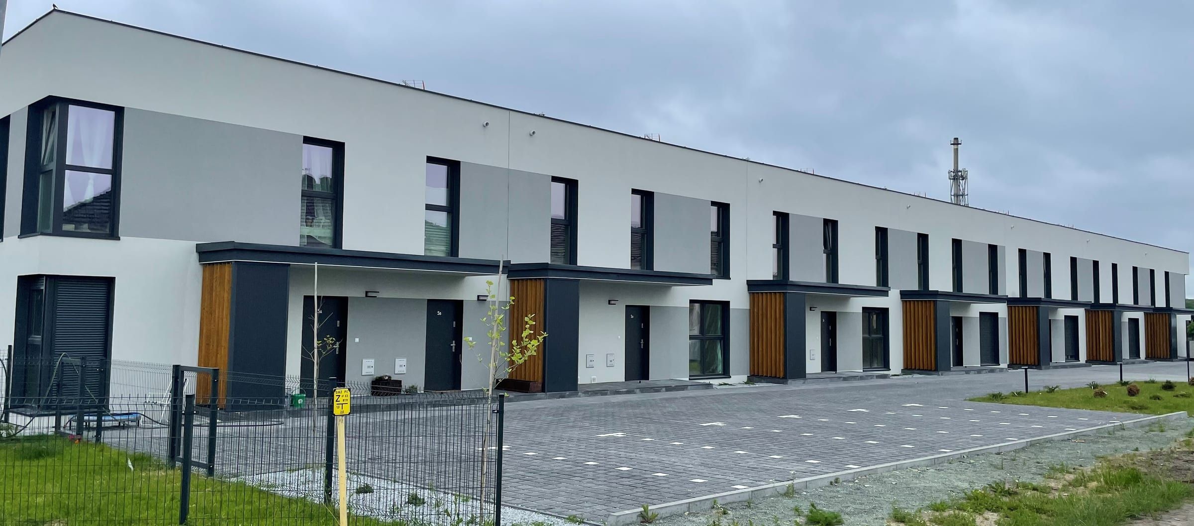 Szeregowy budynek Box House: jasna elewacja z grafitowymi oknami i drewnianymi wnękami wejść, przed nim parking z kostki