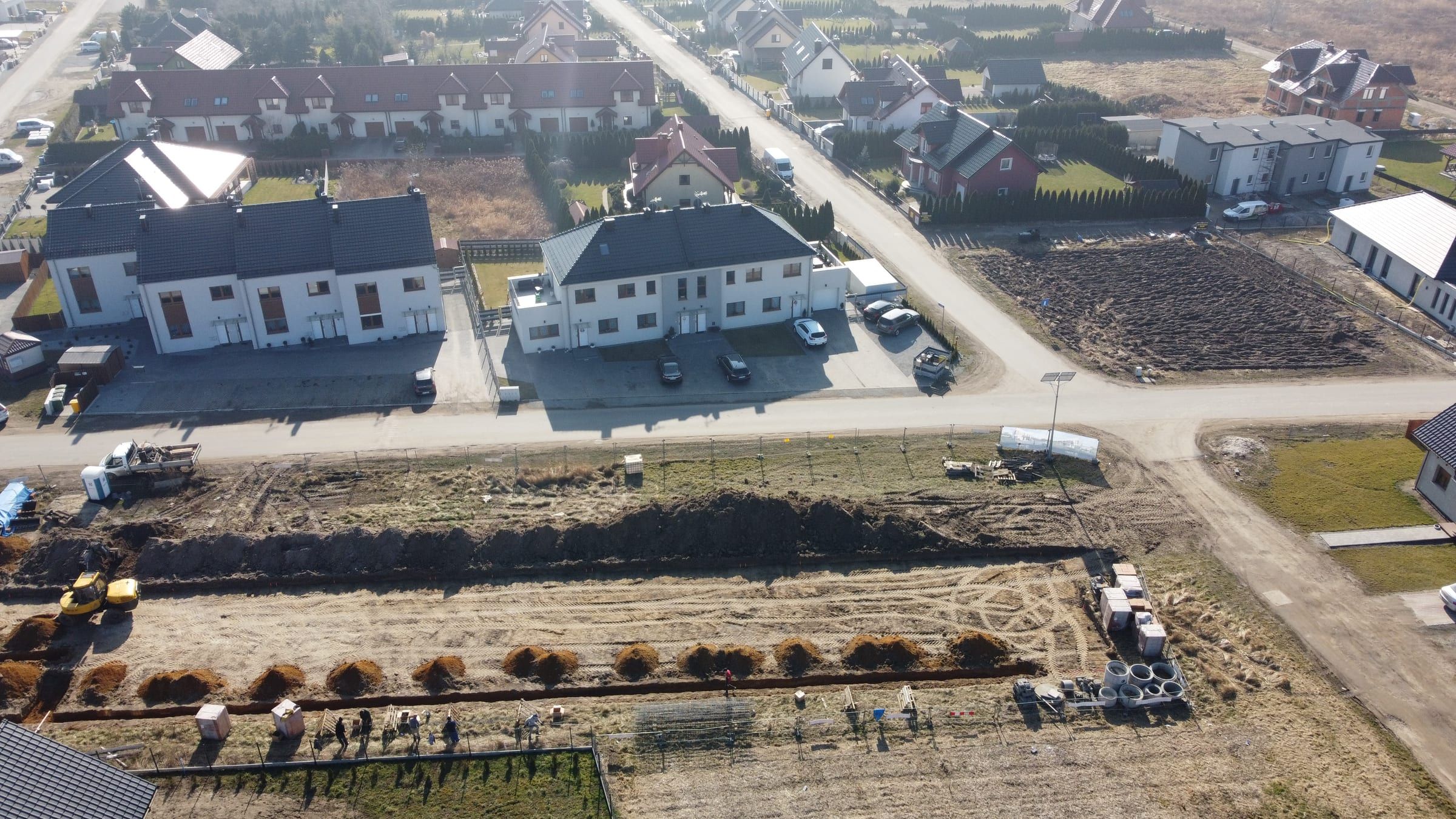 Osiedle z lotu ptaka: białe budynki wielorodzinne z grafitowymi dachami przy osiedlowej ulicy, za nimi szeregowiec z czerwonym dachem i domy jednorodzinne, na pierwszym planie wykopy pod fundamenty nowej budowy